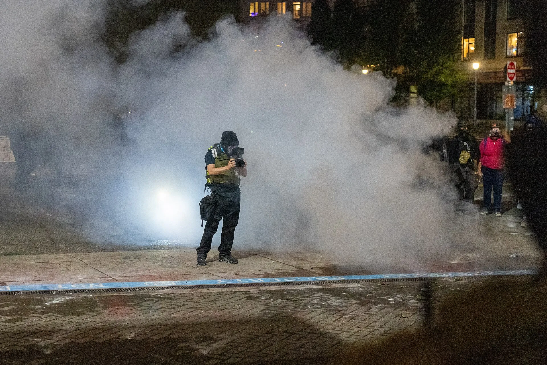 A journalist in a helmet and gas mask stands in a thick cloud of white gas outside the ICE facility in Portland at night.